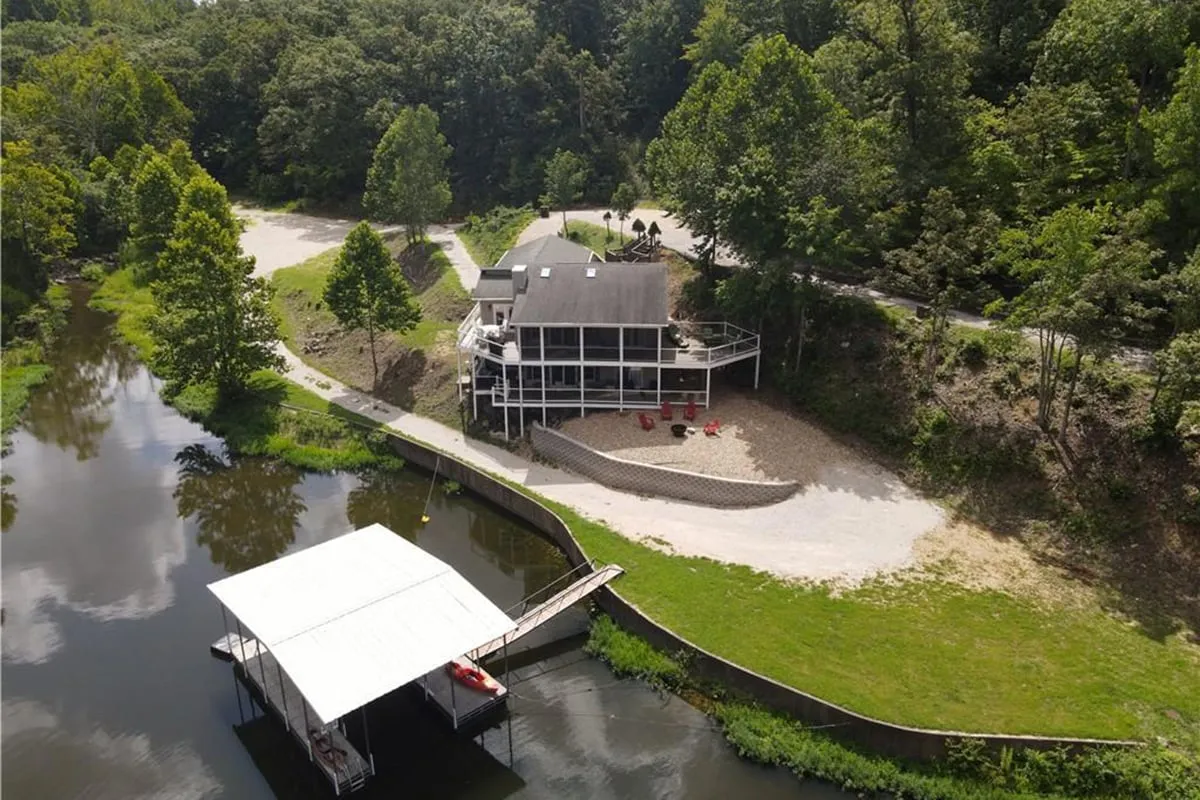 The image showcases a beautifully updated single-family house nestled among mature trees, overlooking a tranquil private cove on Lake of the Ozarks. The house features a spacious deck perfect for sunset views, a flat green lawn leading to a private dock, and calm waters ideal for boating, fishing, or relaxing. The dock and boat slip are visible, hinting at lakeside adventures, while the peaceful surroundings evoke a sense of relaxation and escape.