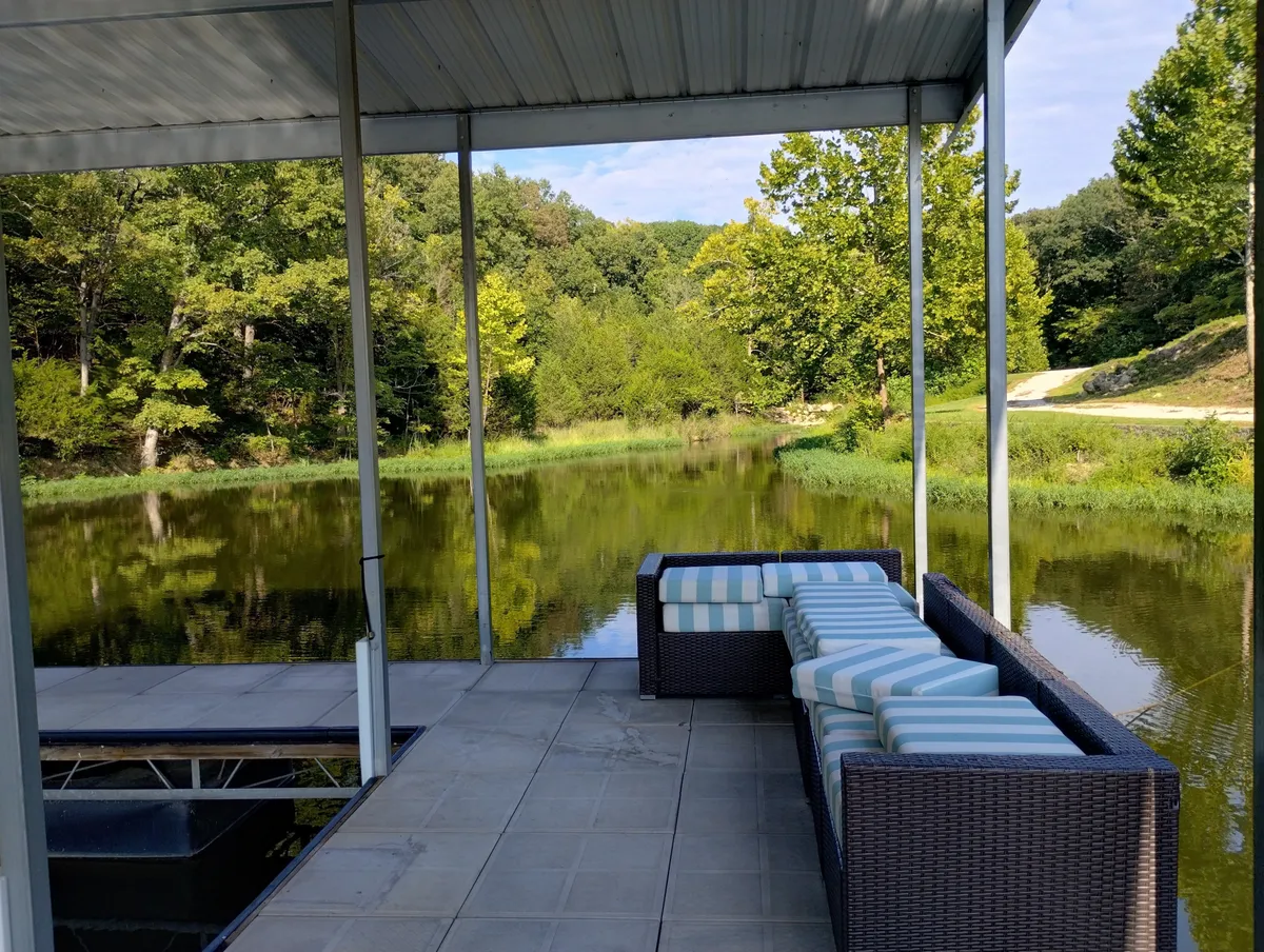 This image captures an outdoor seating area situated beside a reflective body of water, surrounded by lush greenery. The tranquil setting is ideal for relaxing or gathering with friends after an evening out, and the soft lighting suggests the onset of sunset or early evening.