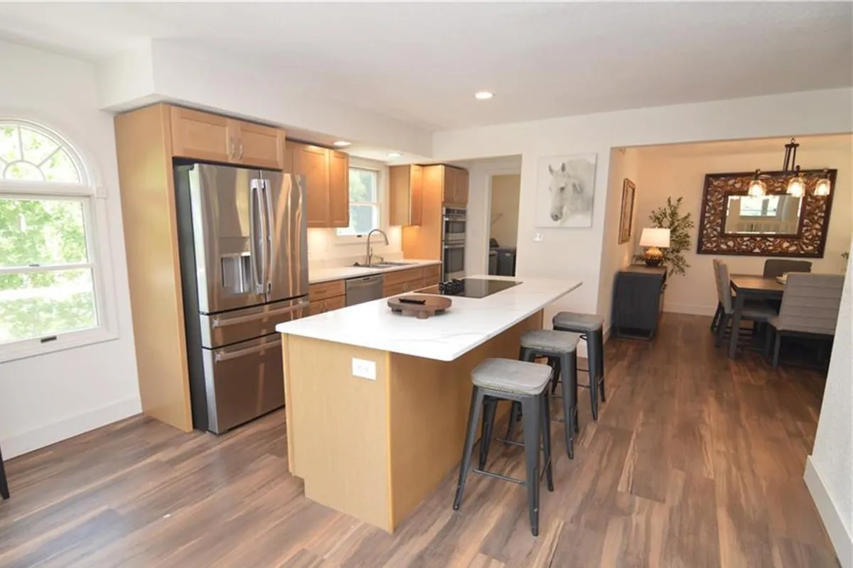 The image features a welcoming kitchen with a prominent island, stainless steel appliances, a dining area, and large windows that bathe the room in natural light. It embodies a space for cooking, entertaining, and family gatherings, with a seamless flow between kitchen and dining zones.