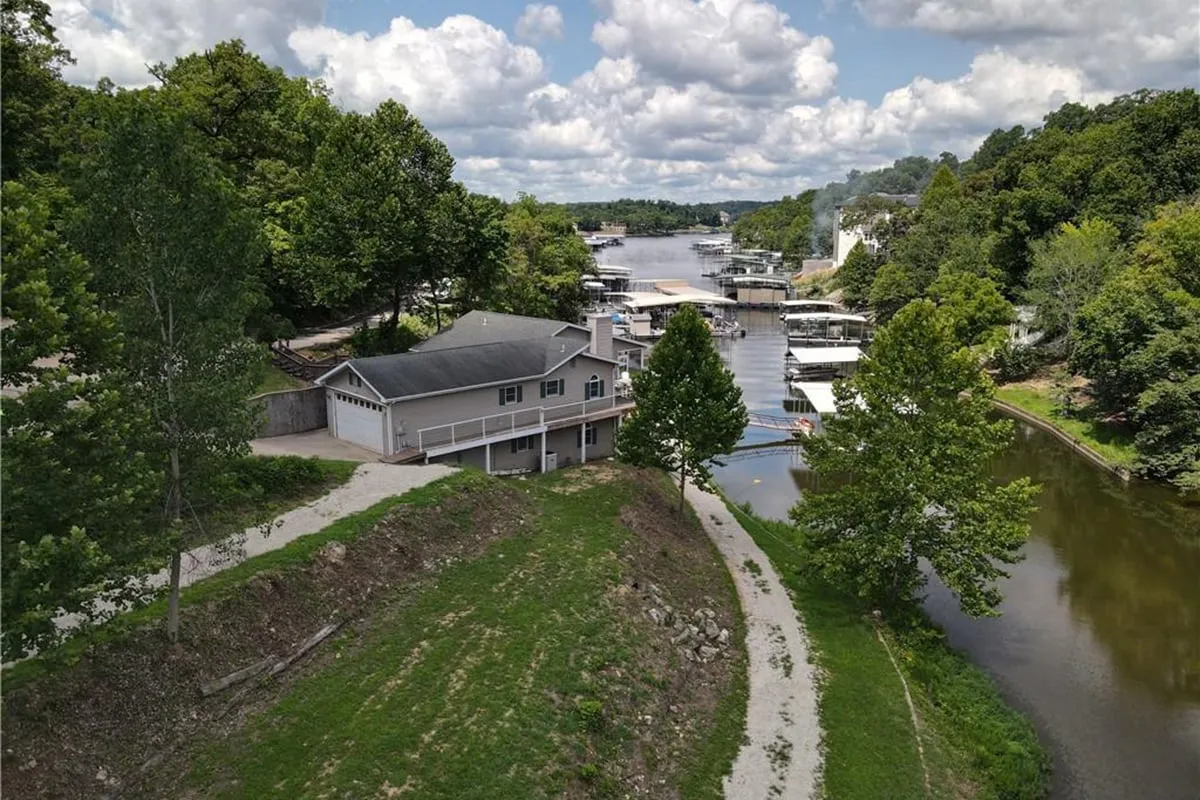 A broad exterior view that includes the house, marina with boat slips, a winding path, and surrounding trees, all set alongside the water. The perspective showcases both the property’s proximity to the lake and the accessibility of boating facilities.