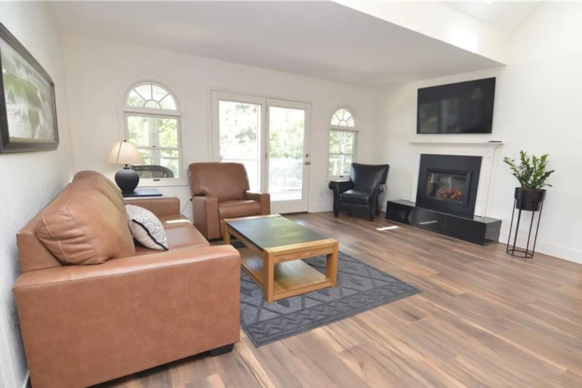 This cozy living room scene features leather sofas, a fireplace, a coffee table, and expansive windows allowing natural light and lake views to filter in. The space is perfect for relaxing, reading, or sharing coffee with friends and family.