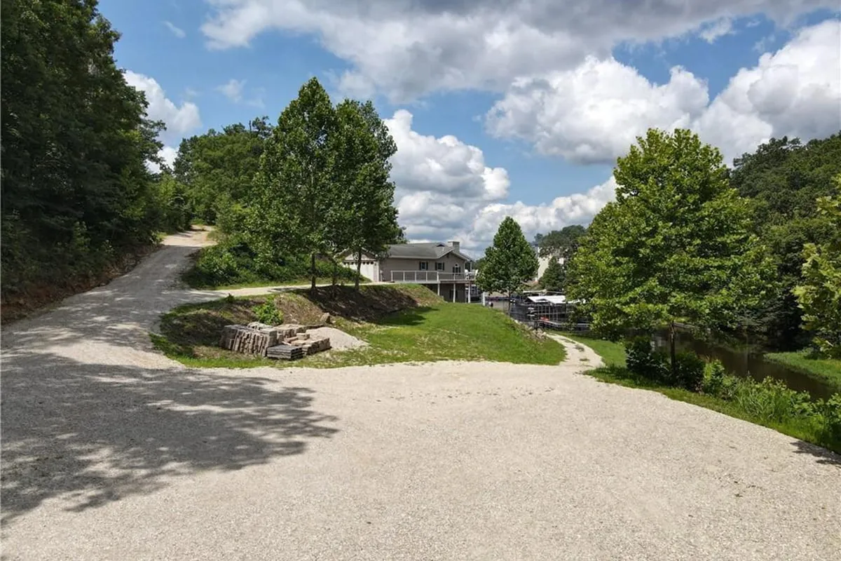 An inviting exterior shot showcasing a gravel driveway leading to the house, framed by lush green trees and a tranquil pond on the right. The peaceful backdrop highlights the natural beauty surrounding the property and hints at outdoor gatherings or quiet evenings under the stars.