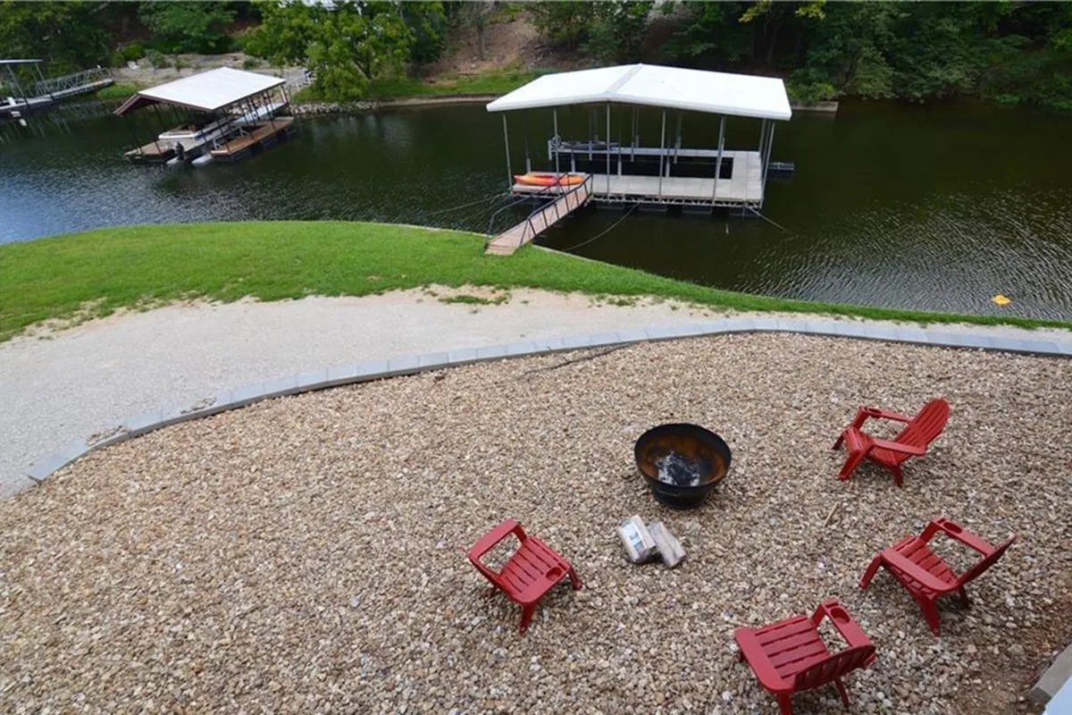 A cozy lakeside scene with a fire pit surrounded by red chairs, with the dock and water visible in the background. The setting suggests evening gatherings, boating, fishing, and relaxing by the water, all in a communal, inviting atmosphere.