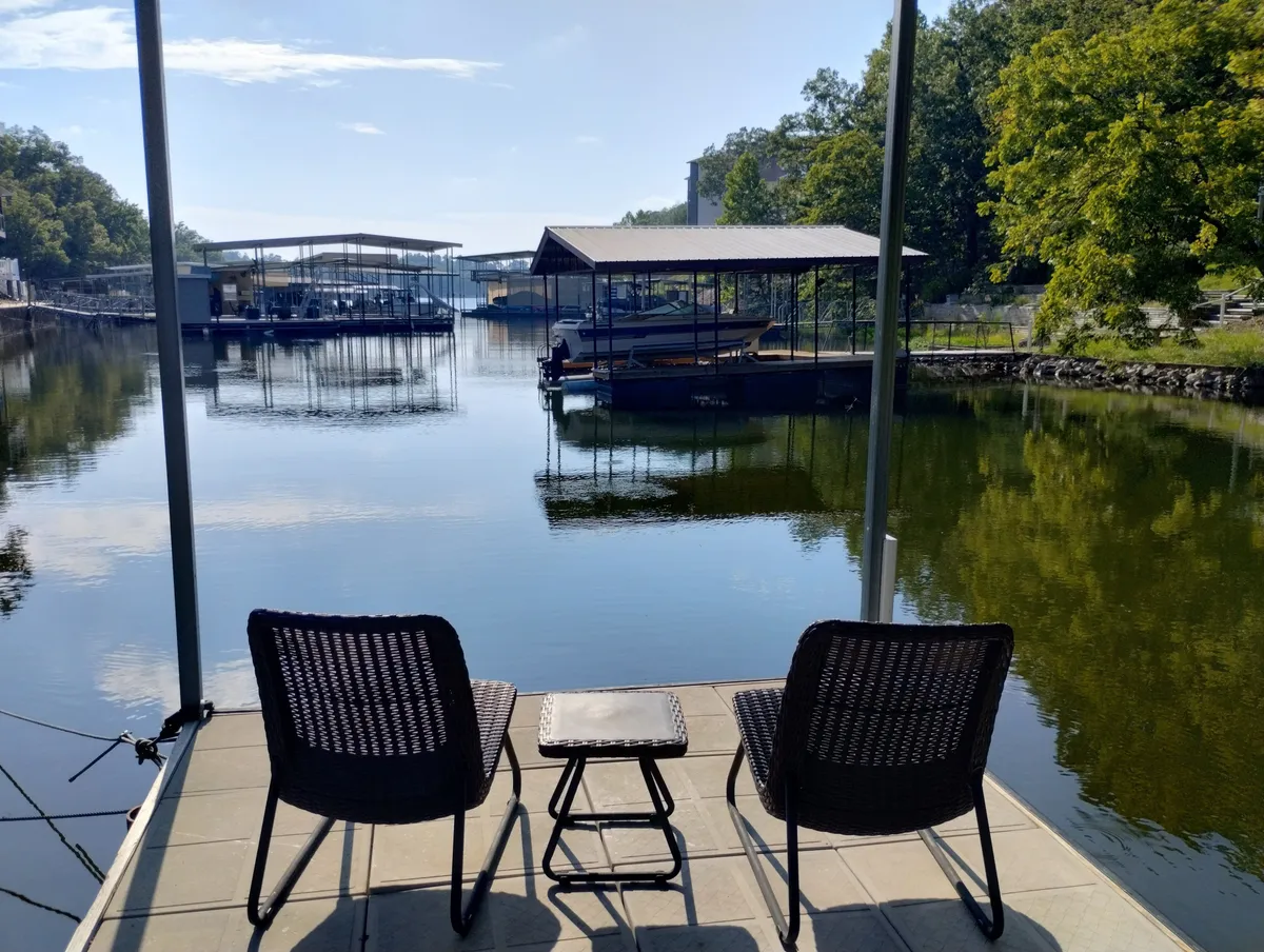 Two black wicker chairs and a small table set on a peaceful dock overlooking calm water, with boats visible in the background and trees lining the shore. The scene is serene, with a focus on outdoor relaxation, enjoying coffee, or watching boats drift by.