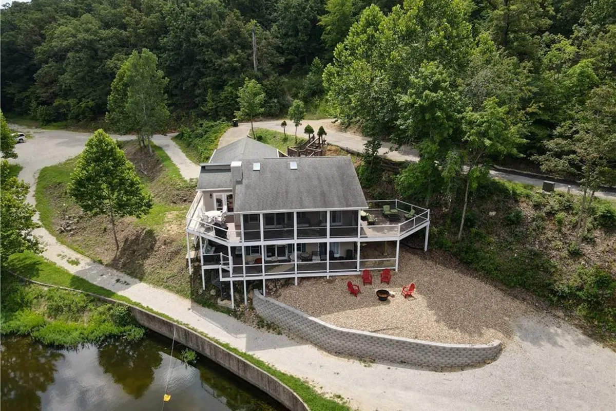 This exterior image showcases the house set against a waterfront backdrop, with red chairs arranged for outdoor seating, mature trees providing shade, and a welcoming atmosphere perfect for group gatherings. The fire pit area and proximity to the water highlight spaces for evening relaxation and socialization.